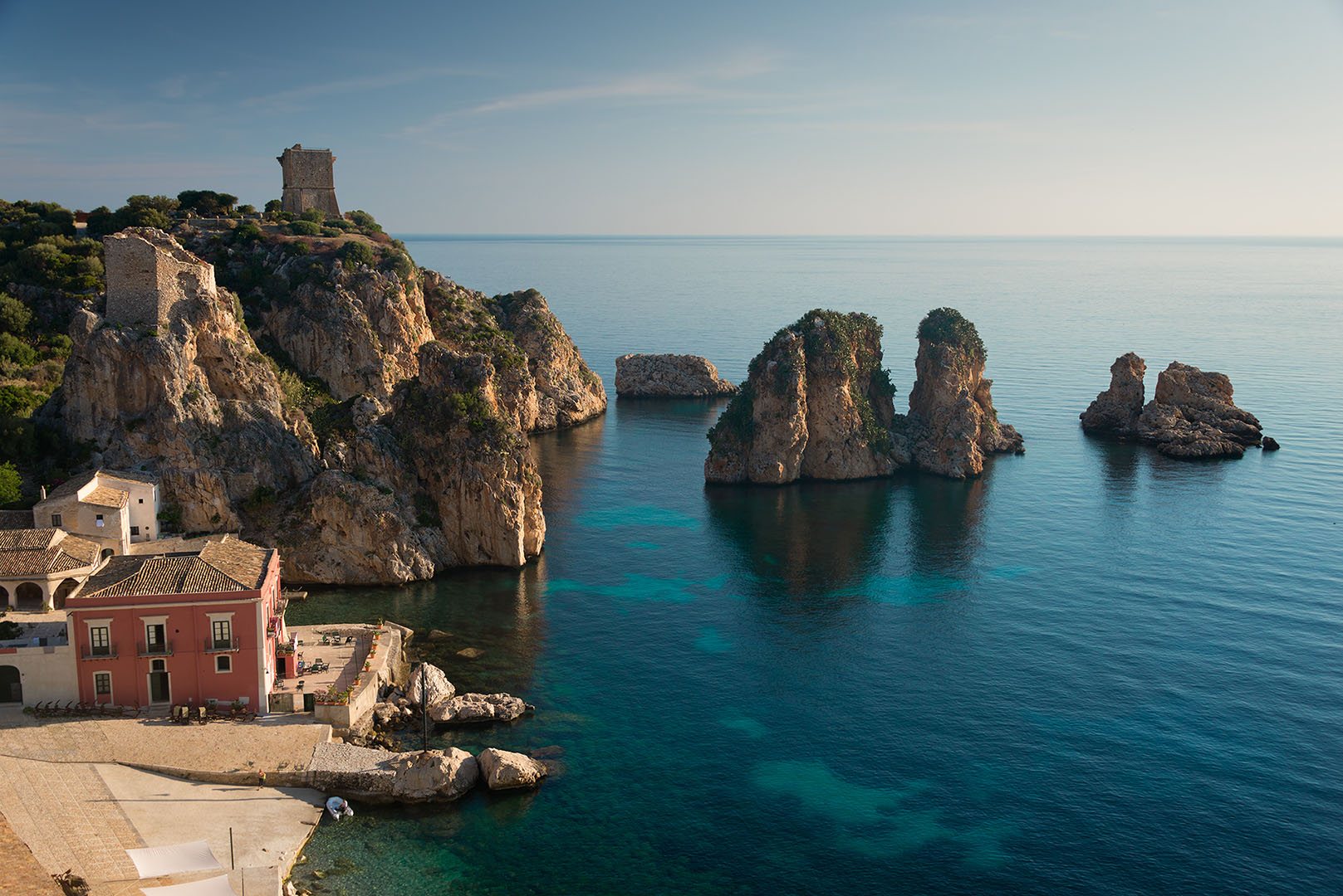 The I Faraglioni rocks in the Mediterranean Sea and the old red Tonnara building in the early morning sunlight in the town of Scopello, at the foot of the Lo Zingaro Nature Reserve at the northern coast of the island of Sicily, Italy.